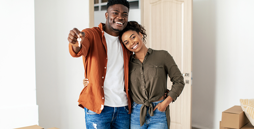 A couple standing in the doorway of their new home, one of them holding up the keys, with moving boxes behind them.
