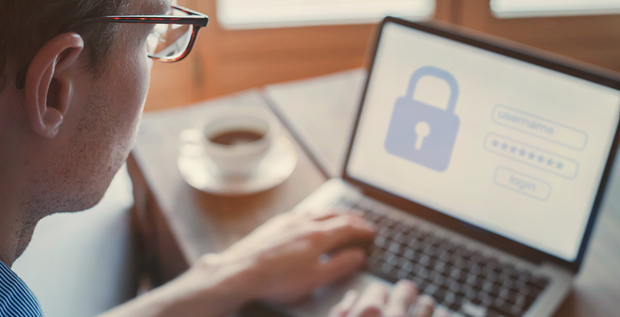 A man at a laptop showing a login screen with a padlock icon, a coffee cup beside him.