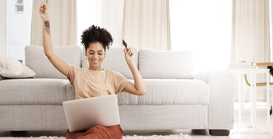 A woman sitting on the floor beside a sofa with a laptop, raising her arms in celebration while holding a card.