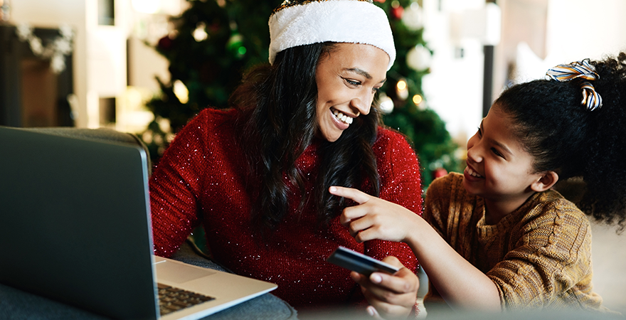 A woman in a Santa hat and a child shopping online at a laptop with a bank card, a Christmas tree behind them.
