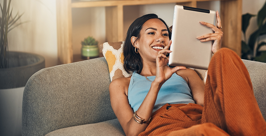 A woman relaxing on a couch, smiling at the tablet she is holding.