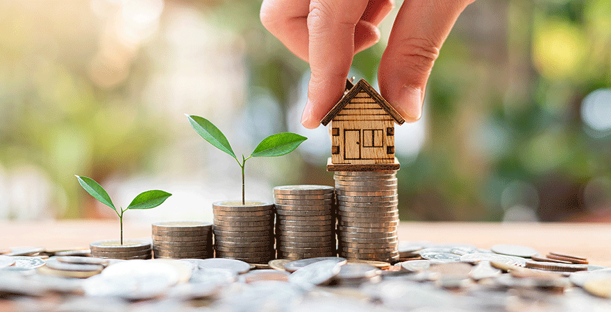 A small wooden house model resting on the tallest of several rising stacks of coins with seedlings growing from them.