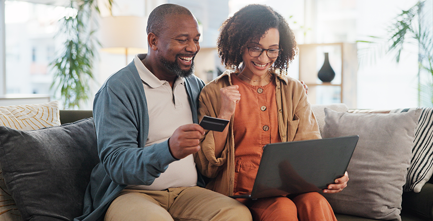A couple sitting together on a sofa with a laptop, one of them holding a bank card.