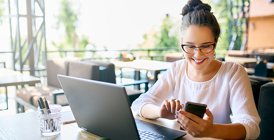 A woman working at a laptop in a cafe while holding her phone.