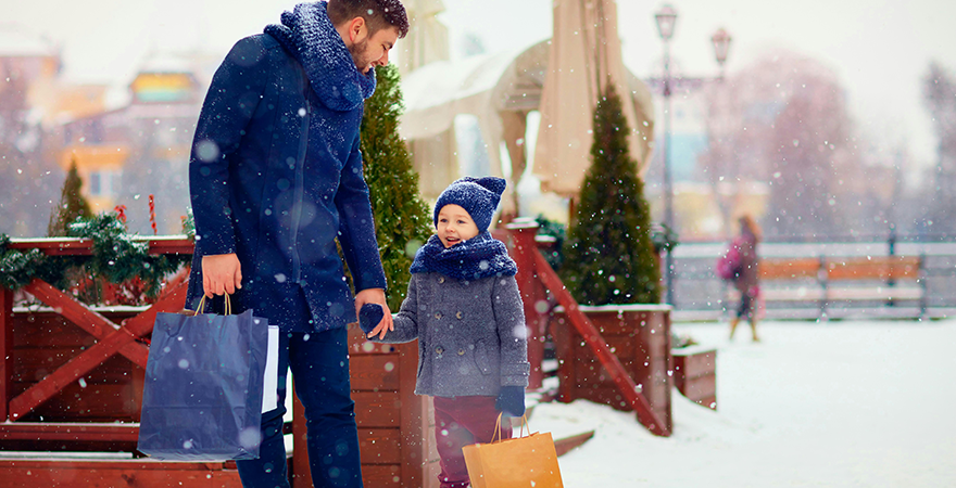 An adult and a young child carrying shopping bags through a snowy town square.