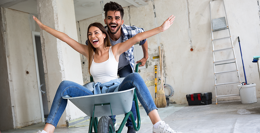 A smiling couple in a room under renovation, one pushing the other in a wheelbarrow.