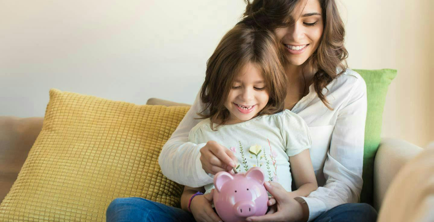 A parent and young child putting a coin into a pink piggy bank on a sofa.