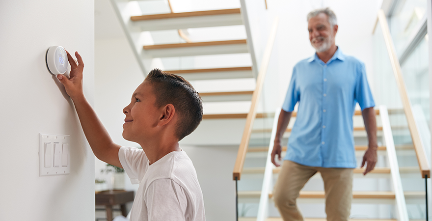 A child adjusting a thermostat on a hallway wall while an adult walks down the stairs behind them.